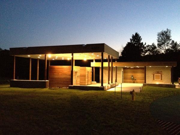 A modern building structure with a wooden façade, illuminated by soft lights, set against a twilight sky. The building features a canopy supported by columns and has sections with open space, surrounded by grass and trees in the background. Lebanon Hills mountain bike trail.