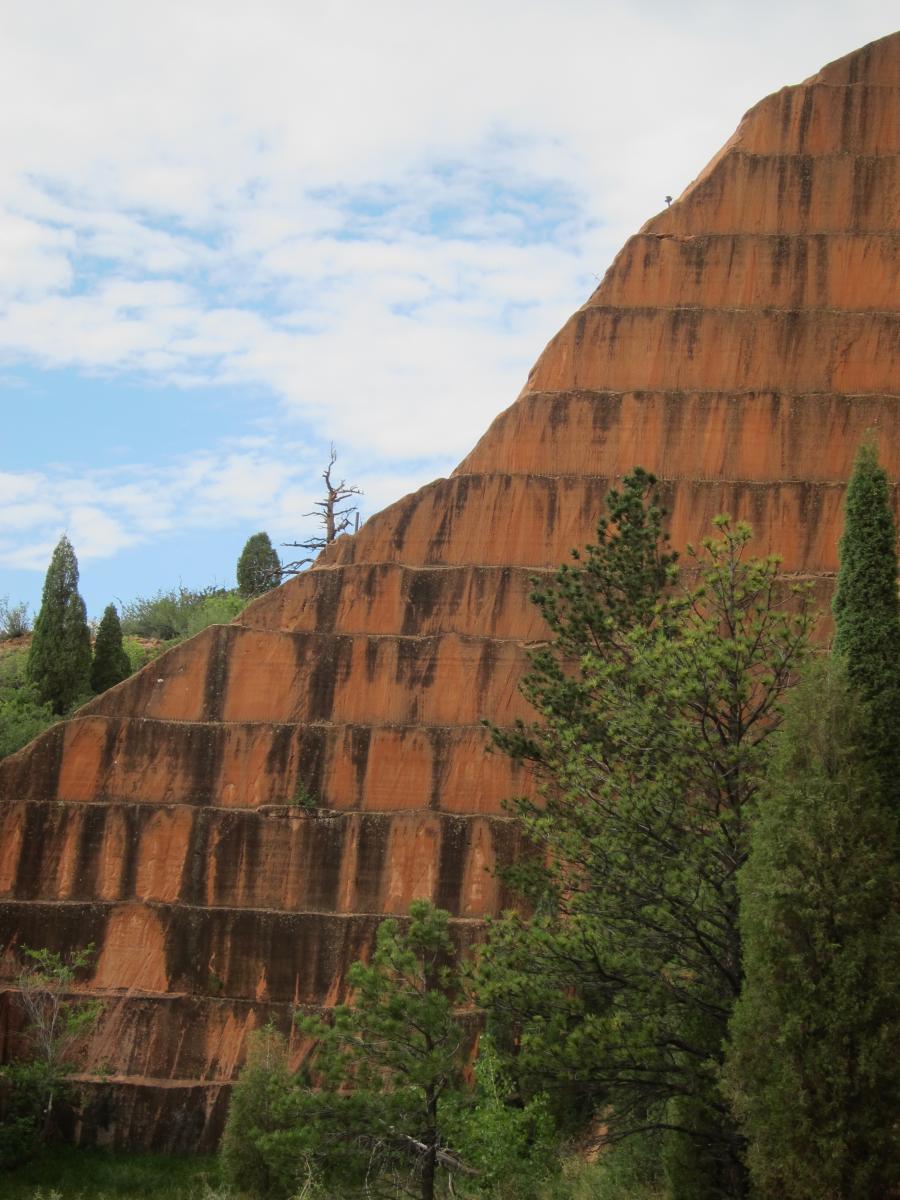A sloping, terraced rock formation with reddish-brown layers, surrounded by green trees and shrubs. The sky is partly cloudy, adding a serene backdrop to the landscape. Red Rock Canyon mountain bike trail.