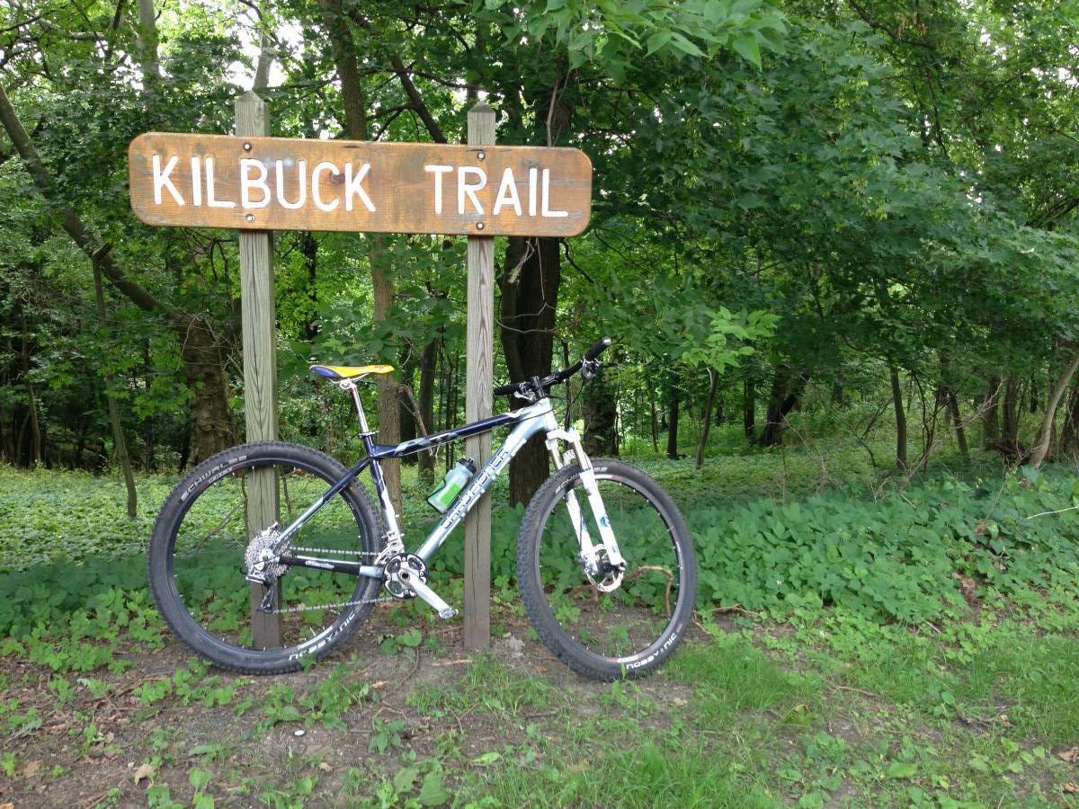 Sign for Kilbuck Trail with a mountain bike leaning against it, surrounded by lush greenery and trees. Riverview Park mountain bike trail.