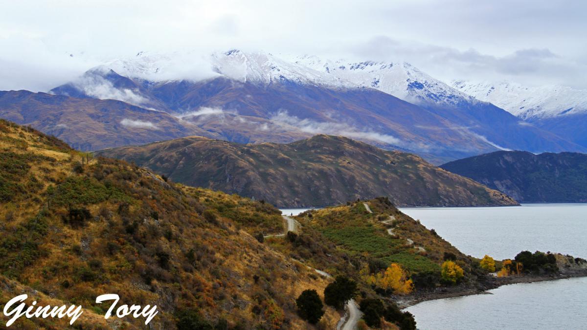 A scenic landscape featuring rolling hills and mountains covered in snow, under a cloudy sky. In the foreground, a winding path runs alongside a body of water, while trees with autumn foliage add splashes of color. The distant mountains provide a dramatic backdrop, contributing to the tranquil beauty of the scene. Glendhu Bay Track mountain bike trail.