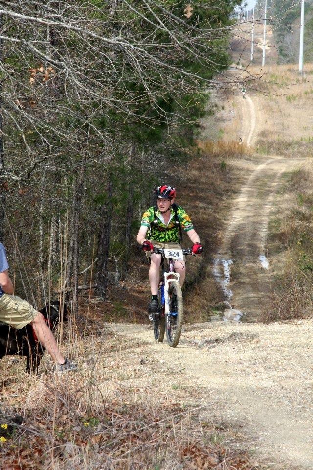 A cyclist wearing a colorful jersey and helmet rides a mountain bike along a dirt trail surrounded by trees. In the foreground, a person seated on the ground with a dog is visible, while the path continues into the background, leading into a grassy area. Arrowhead Park mountain bike trail.