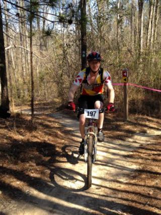 A mountain biker wearing a jersey with red and yellow accents rides on a dirt trail through a wooded area. The biker has a number pinned to their jersey (197) and is approaching a trail marker. Sunlight filters through the trees, casting shadows on the path. Arrowhead Park mountain bike trail.