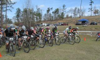 A group of mountain bikers at the start line of a race, with several cyclists in motion on their bikes. They are wearing helmets and race numbers, surrounded by a grassy area and trees in the background. A few vehicles and a tent are visible in the distance under a clear blue sky. Arrowhead Park mountain bike trail.