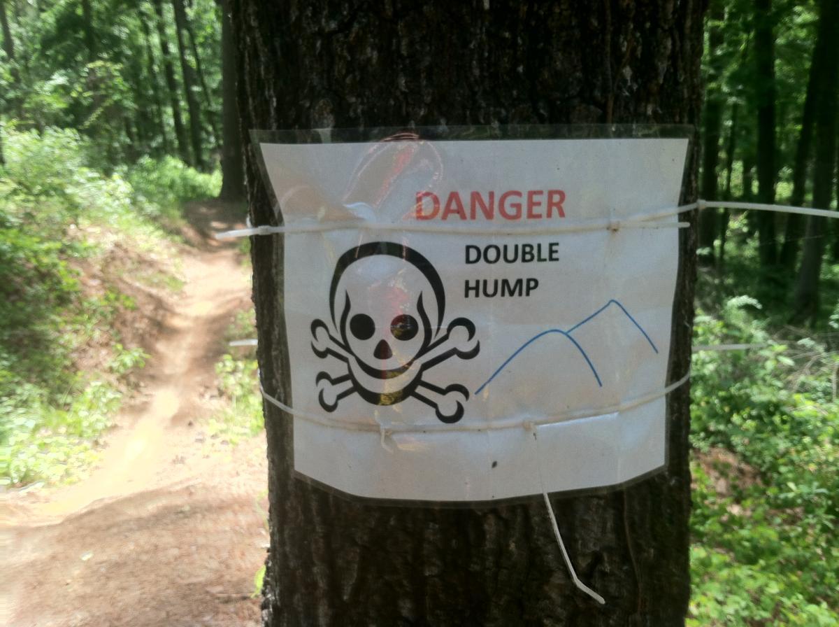 Sign on a tree warning of danger ahead, featuring a skull and crossbones graphic. The text reads "DANGER DOUBLE HUMP." The background shows a winding dirt path in a forested area. Forks Area Trail System (FATS) mountain bike trail.