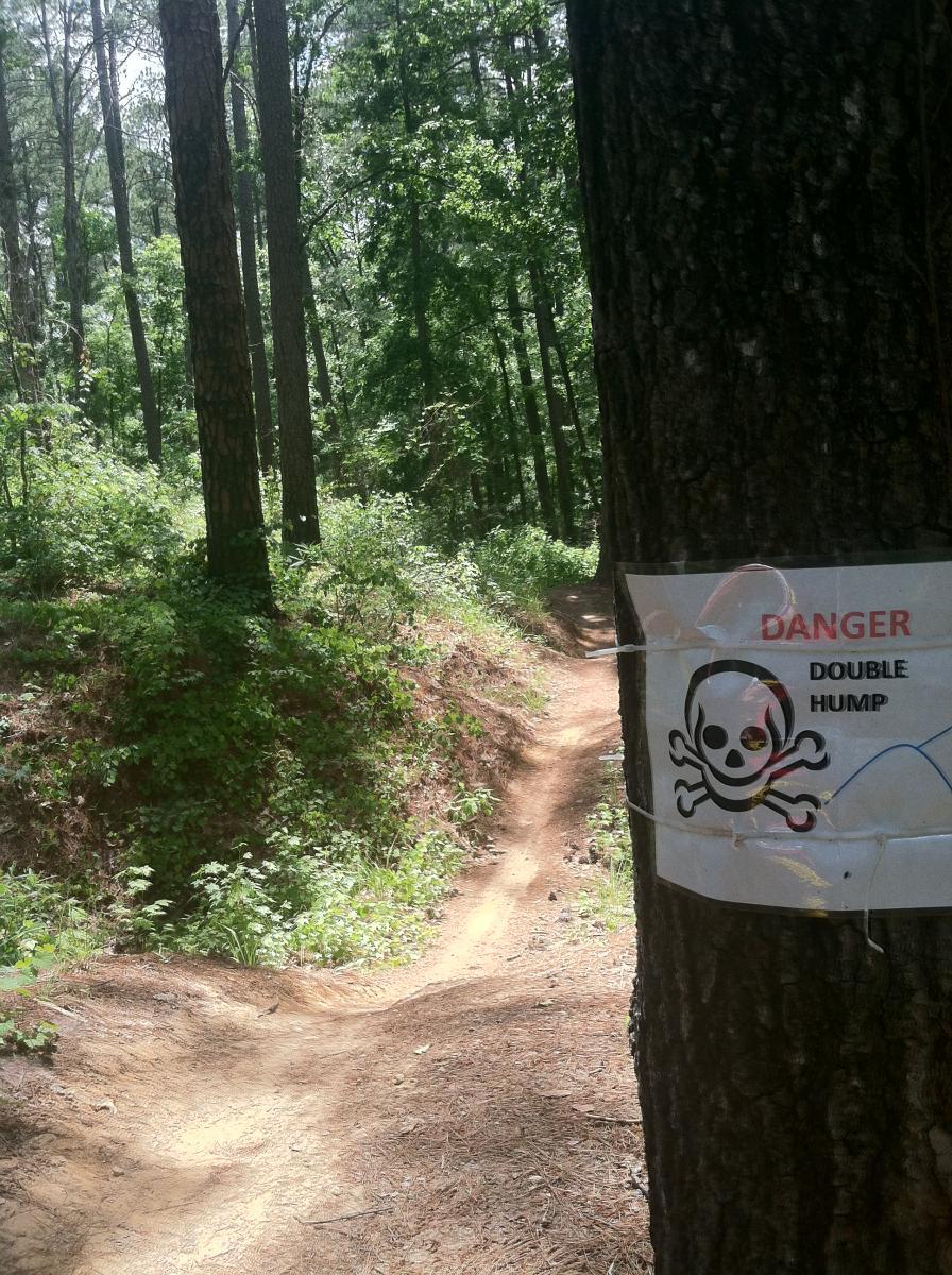 A dirt trail winding through a lush green forest, with a warning sign on a tree displaying "DANGER" and a skull icon, indicating a "DOUBLE HUMP" ahead. Forks Area Trail System (FATS) mountain bike trail.