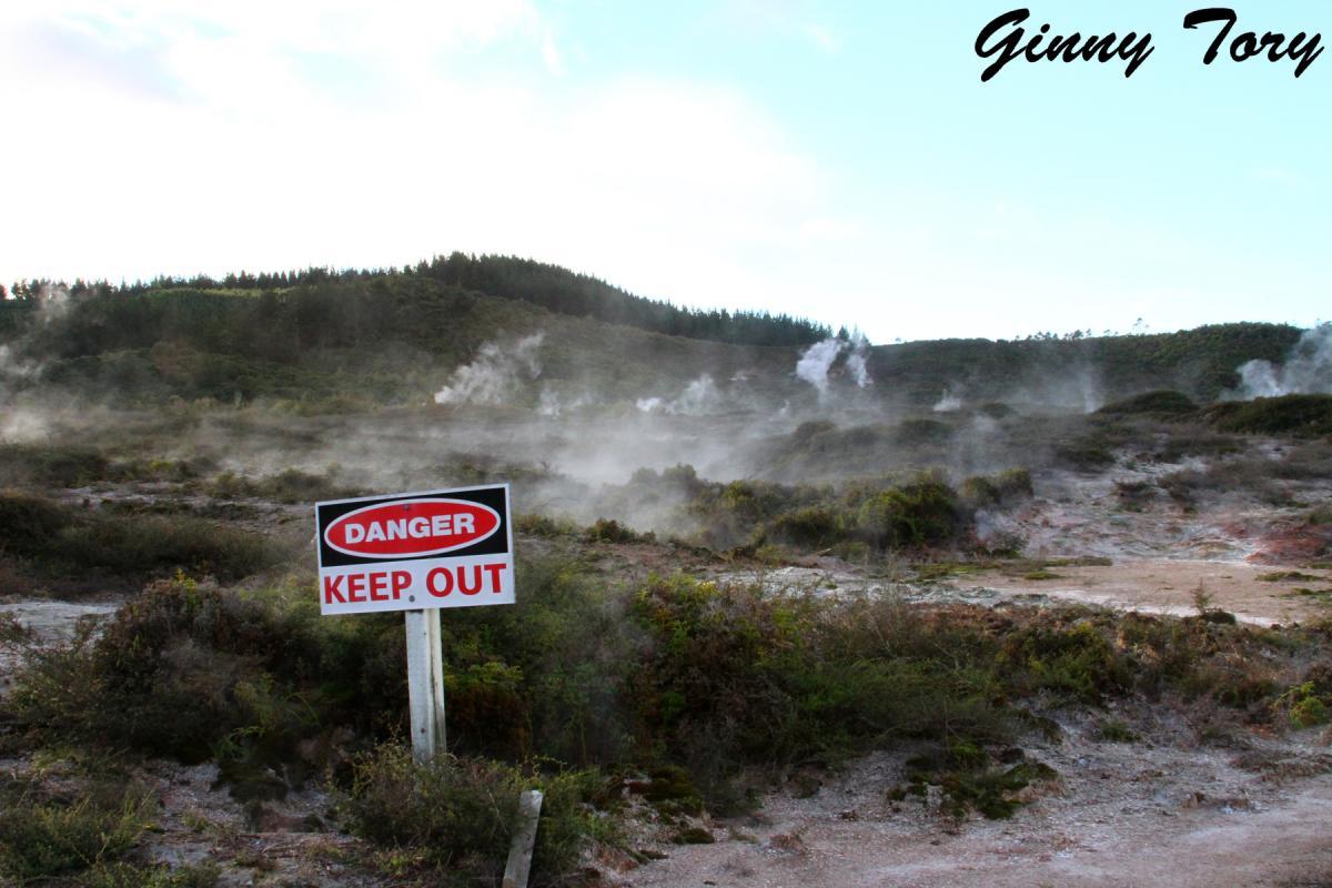 A sign reading "DANGER KEEP OUT" stands prominently in the foreground, alerting to hazardous conditions in the area. Behind the sign, a landscape is shrouded in wisps of steam, with a hilly terrain and patches of vegetation. The sky is partly cloudy, providing a contrast to the earthy tones of the ground. Craters Mountain Bike Park mountain bike trail.