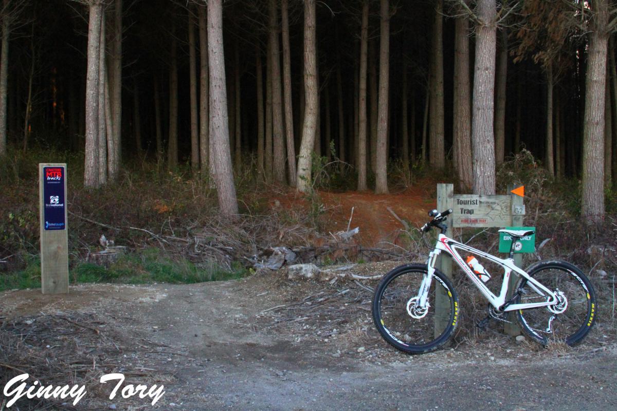 A mountain bike rests against a wooden sign at the entrance to a forest trail. The sign indicates the trail name "Tourist Trap" and has a marker for bike use only. The background shows tall trees and a natural, wooded landscape, suggesting a peaceful outdoor setting. Craters Mountain Bike Park mountain bike trail.