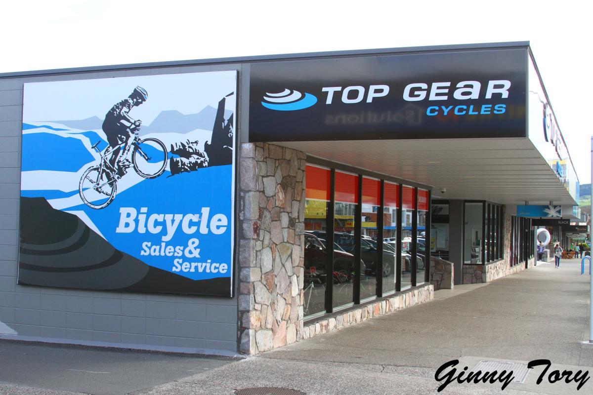 Signage for "Top Gear Cycles," a bicycle sales and service shop, featuring a graphic of a cyclist riding a mountain bike against a blue and white background. The shop has a modern facade with large windows and a stone accent at the entrance.