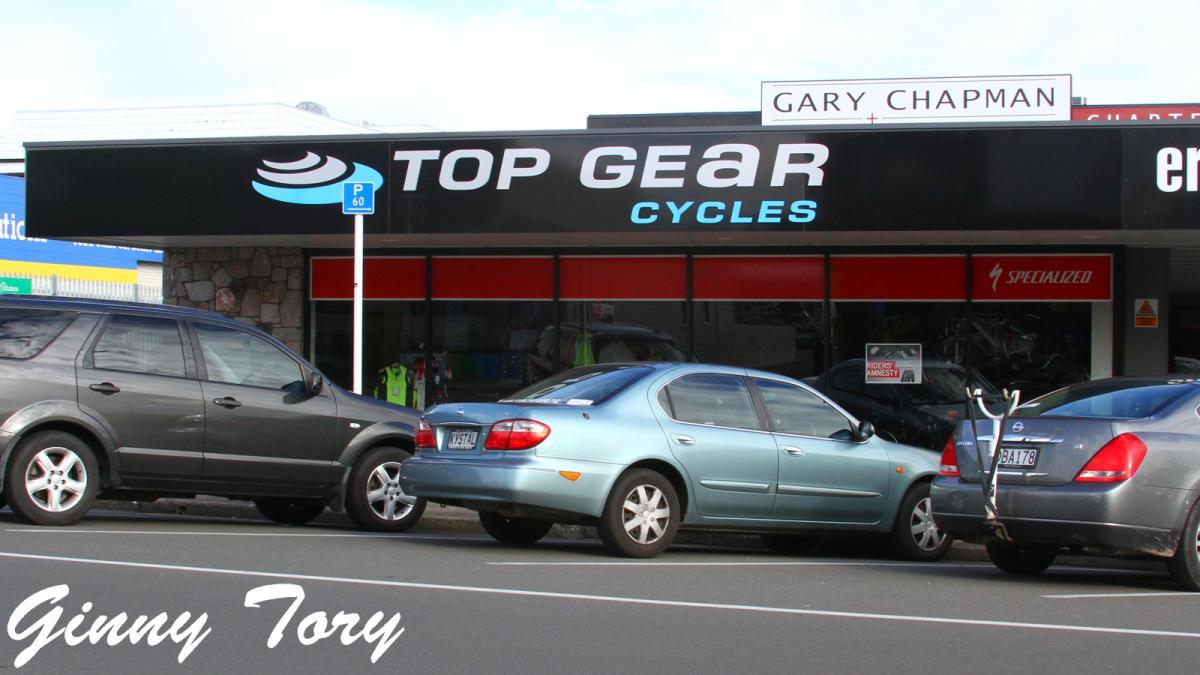 A storefront view of "Top Gear Cycles," featuring bicycles and cycling gear on display inside. The sign prominently displays the store name in bold letters. Several parked cars are in the foreground, with a parking sign visible nearby. The scene is bright and captures an outdoor shopping area.