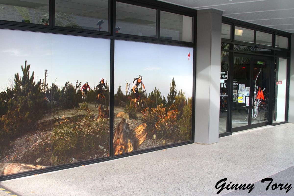 A vibrant mural on the exterior of a shop that depicts mountain bikers riding on a trail surrounded by trees and shrubbery, showcasing an adventurous outdoor scene. The wall features a realistic landscape with clear skies and hints of earthy tones, enhancing the outdoor sports theme.