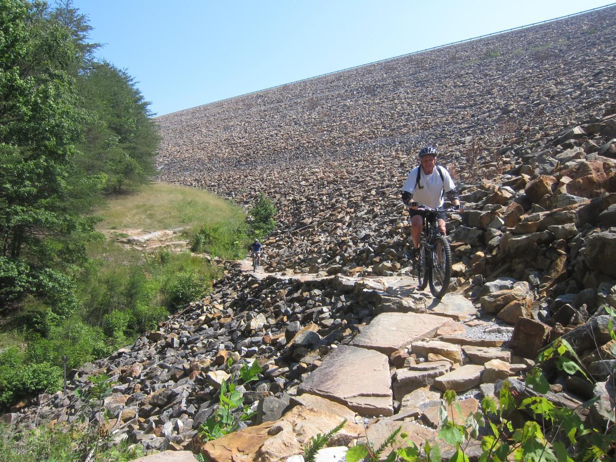 A mountain biker navigates a rocky trail alongside a large earthen dam under a clear blue sky. The path is uneven with stones, and there are trees lining the area. Another biker can be seen in the distance, showcasing an adventurous outdoor setting. Raccoon Mountain Trail Network mountain bike trail.