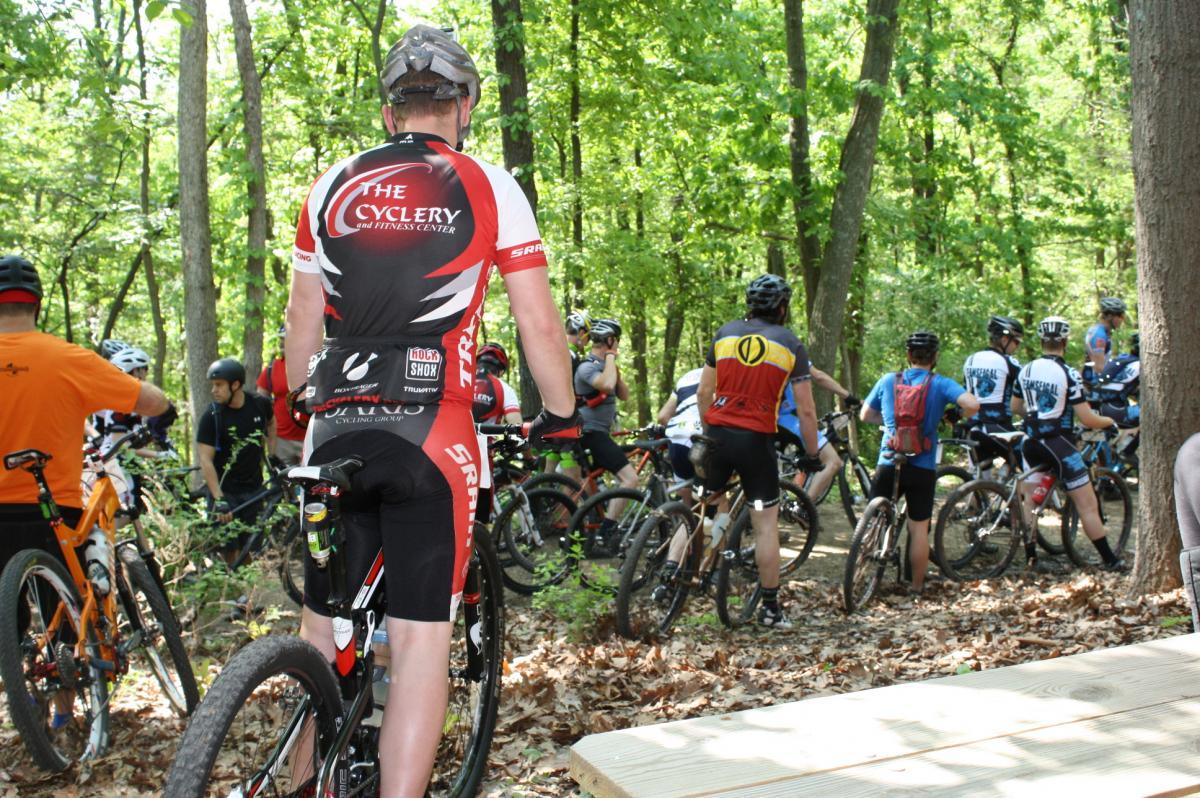 A group of mountain bikers gathered in a forested area, some seated on their bikes and others standing. The cyclist in the foreground is facing away, wearing a red and black jersey with the logo of a cycling center. Surrounding them are various riders in colorful jerseys and helmets, with trees and greenery in the background. Greensfelder Park mountain bike trail.