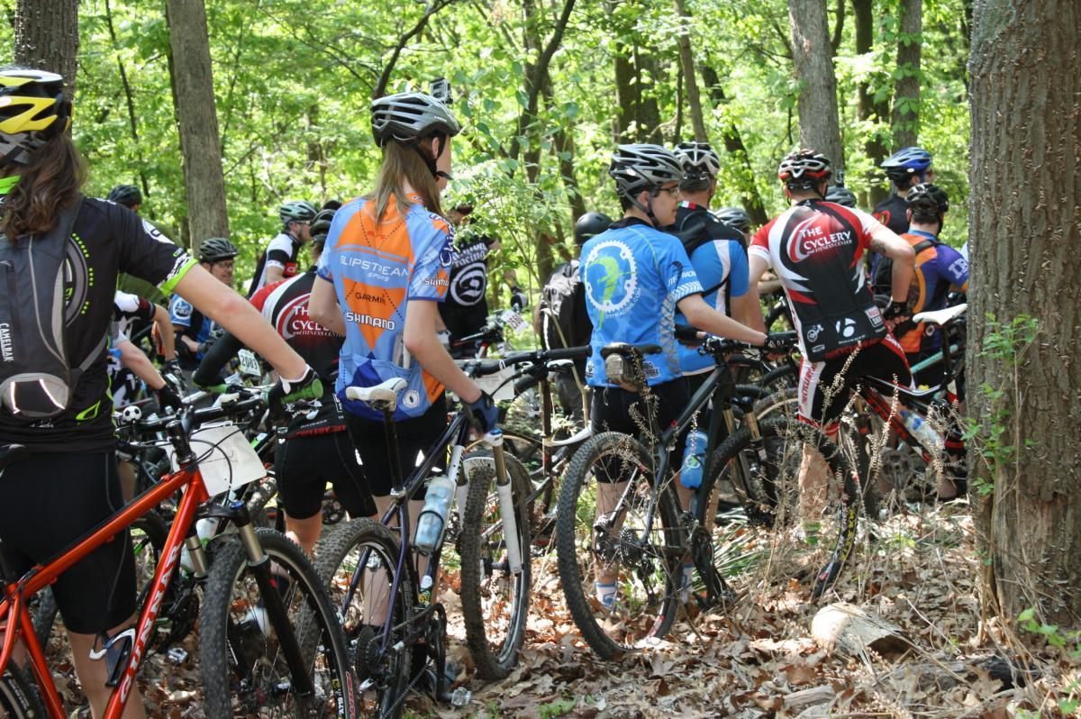 A group of mountain bikers in cycling gear, standing with their bikes in a wooded area. The scene captures participants preparing for a race or event, with trees and foliage surrounding them. Various bike brands and colorful jerseys are visible, indicating a community of cycling enthusiasts. Greensfelder Park mountain bike trail.