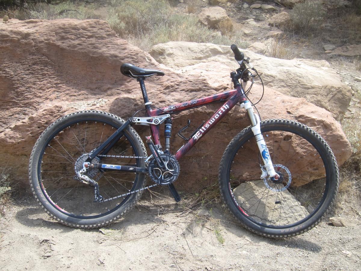 A mountain bike leaning against a large rock in a dirt landscape with sparse vegetation. The bike features a red and black frame, thick tires, and front suspension forks, indicating it is designed for off-road use.