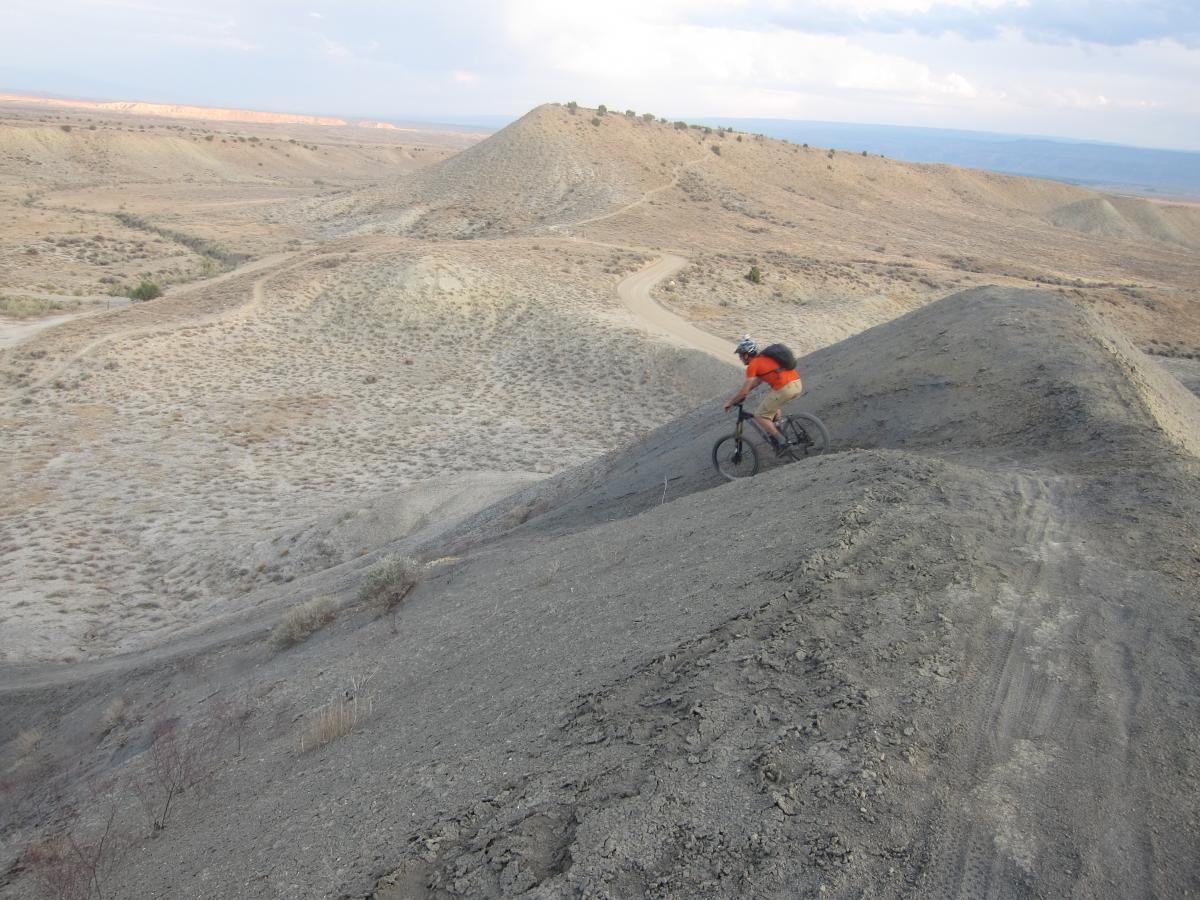 A mountain biker in an orange shirt rides down a steep, rocky hill in a desert landscape, surrounded by rolling hills and a winding dirt path. The terrain is dry and rugged, under a cloudy sky. 18 Road Trails / North Fruita Desert mountain bike trail.