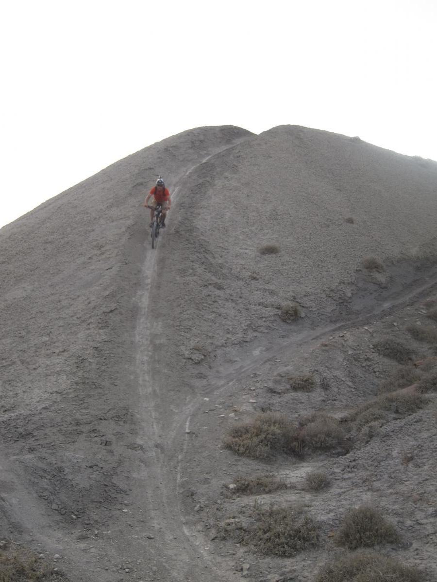 A mountain biker riding along a narrow dirt trail on a steep, rocky hill. The landscape is barren, featuring gray soil and sparse vegetation, with a bright sky in the background. Zippety Do Dah mountain bike trail.