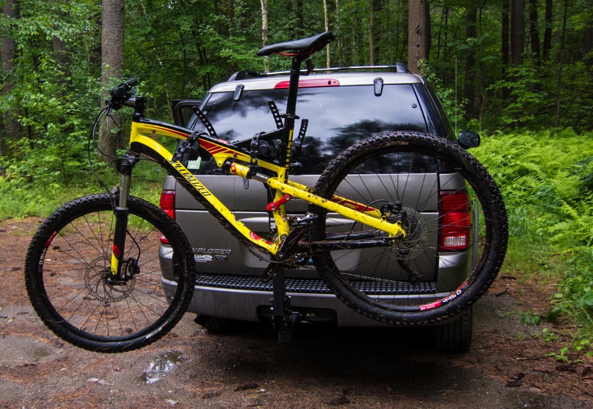 Specialized Camber: A mountain bike mounted on the back of a silver SUV, set against a backdrop of green trees and foliage. The bike features a yellow frame with red accents and has mud on its tires, indicating recent use. The setting appears to be a forested area, with a gravel path visible in the foreground.