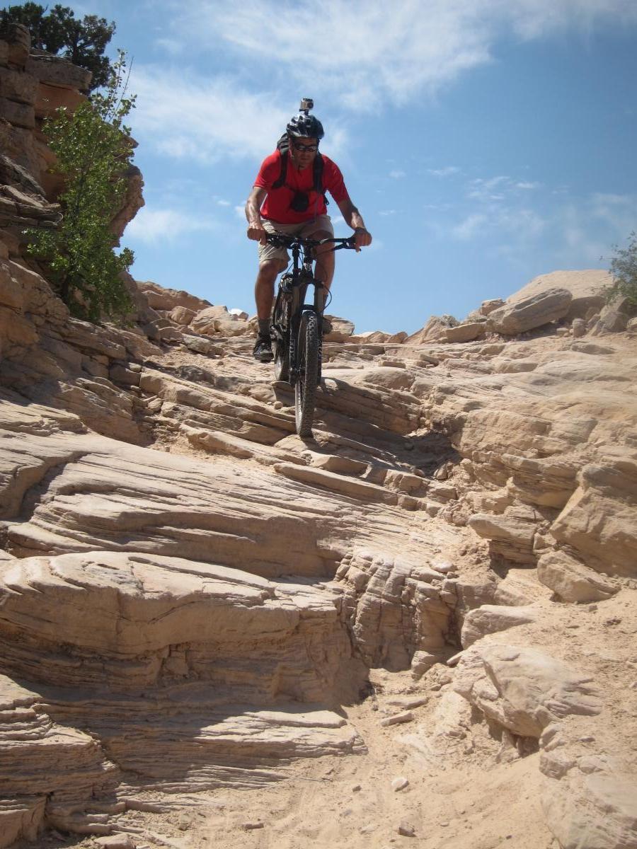 A mountain biker in a red shirt and helmet descends a rocky, sandy trail surrounded by desert landscape under a blue sky. Mary's Loop / Horsethief Bench mountain bike trail.
