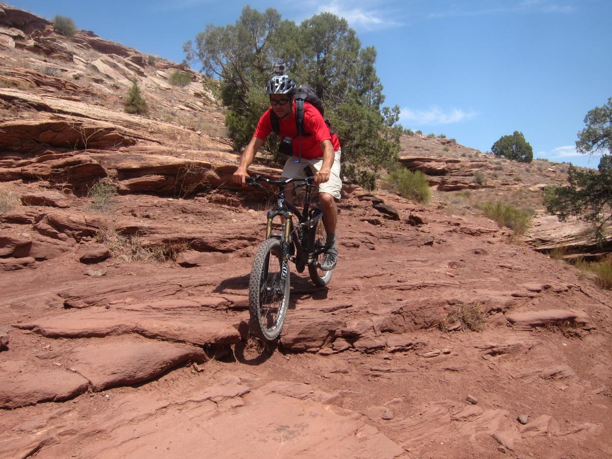 Mountain biker navigating rocky terrain under a clear blue sky, surrounded by desert landscape with trees and sandstone formations. Mary's Loop / Horsethief Bench mountain bike trail.
