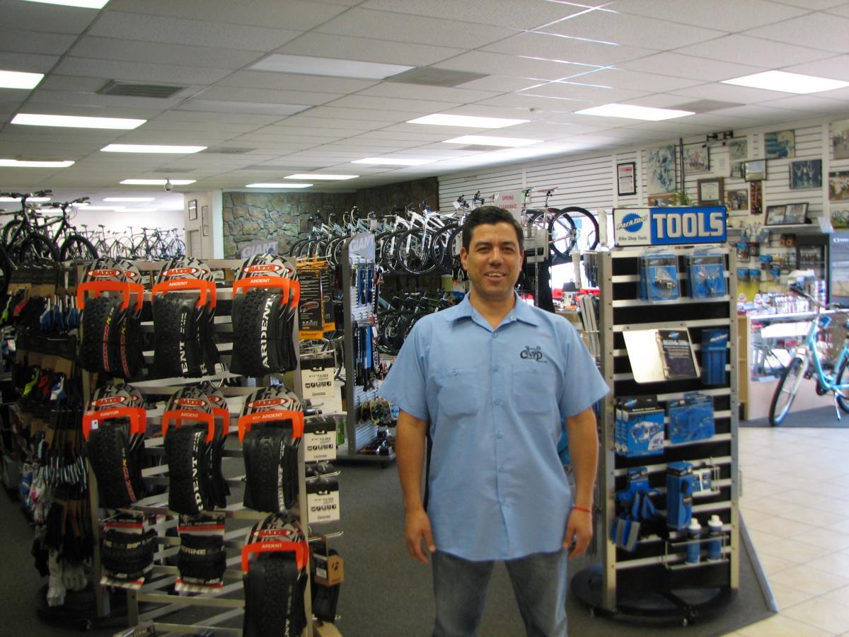 A bicycle shop interior featuring a smiling employee standing in front of shelves displaying bicycle tires and tools. The background includes various bicycles and biking accessories, creating a welcoming environment for customers.