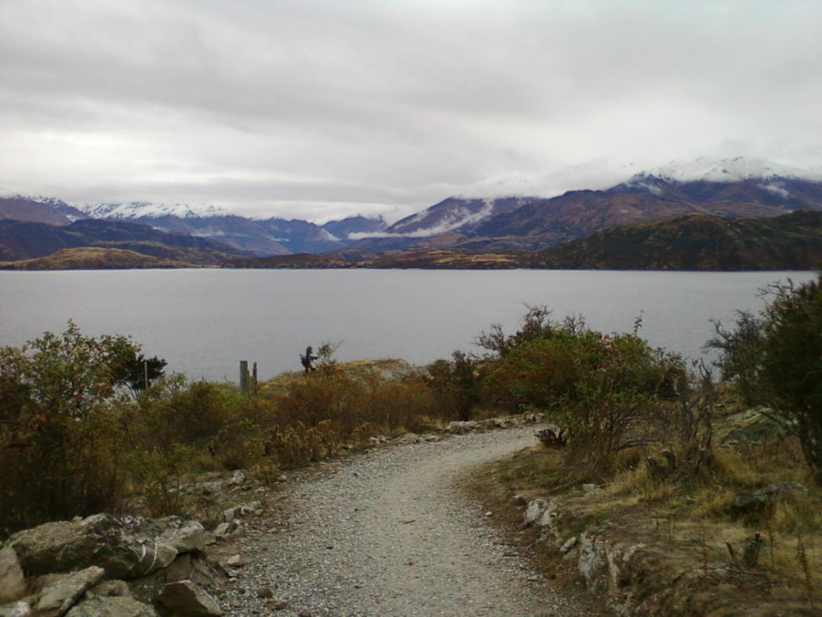 A scenic view of a tranquil lake surrounded by mountains, with a cloudy sky overhead. A gravel path winds along the water's edge, bordered by shrubs and rocky outcrops. Snow-capped peaks are visible in the distance, creating a serene natural landscape. Glendhu Bay Track mountain bike trail.