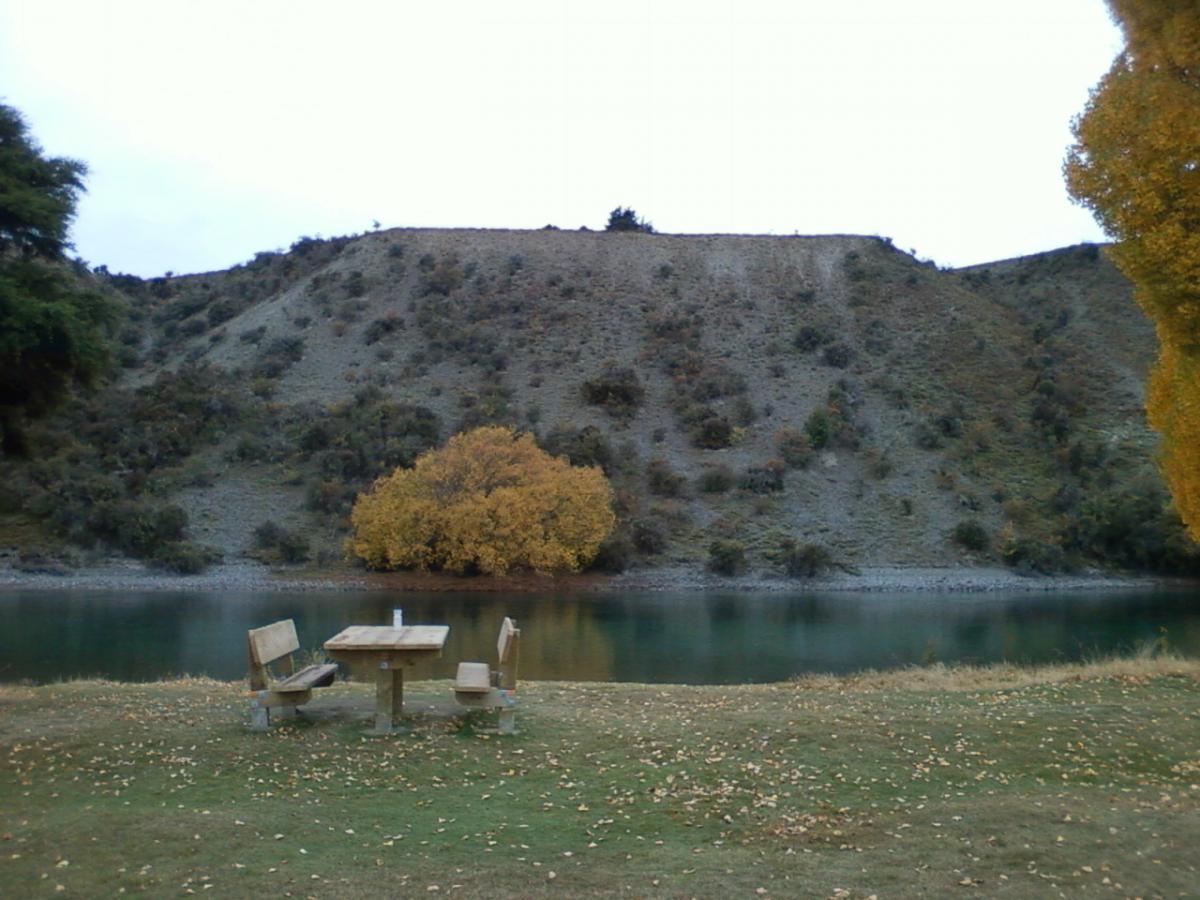 A peaceful outdoor scene featuring a wooden picnic table with two chairs beside a calm river. In the background, a hillside is visible, covered with greenery and dotted with patches of autumn foliage. A large, yellow-leaved tree stands prominently near the water's edge, adding a touch of color to the serene landscape. The sky is overcast, contributing to a tranquil atmosphere. Lake Wanaka Outlet Track mountain bike trail.