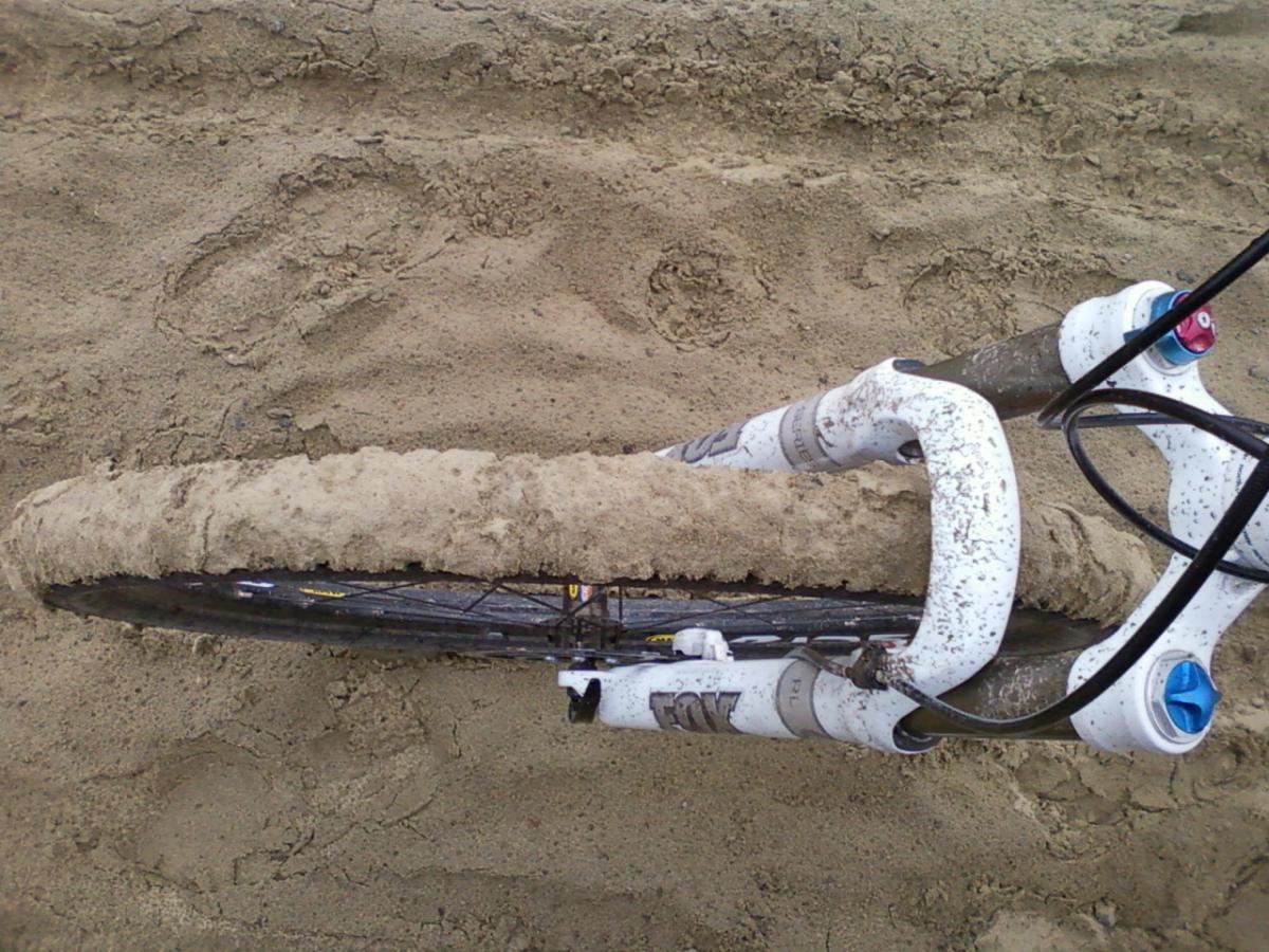A top-down view of a mountain bike partially buried in sand, showcasing a tire covered in sand and dirt. The sandy surface around the bike features footprints and tracks, indicating recent activity in the area. Lake Wanaka Outlet Track mountain bike trail.
