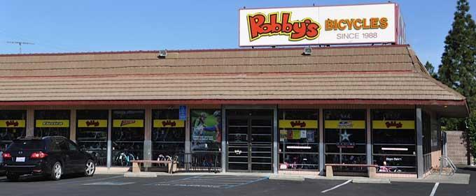 Front view of Robby's Bicycles store, featuring a red and yellow sign with the store name and the text "Since 1988." The shop has large windows displaying various bicycles, and there is a parking lot with a black car in front.