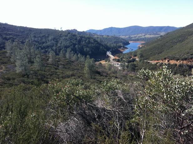 A panoramic view of a lush green valley with rolling hills and a winding river. In the foreground, there are shrubs and trees, while the background features a serene water body and distant mountains under a clear blue sky. Sweetwater Trail mountain bike trail.