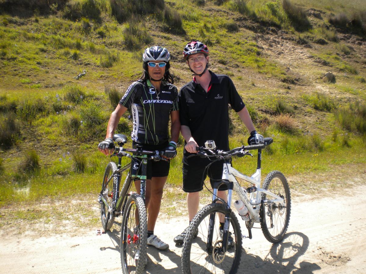 Two mountain bikers pose together on a dirt trail surrounded by grassy hills. One rider, wearing sunglasses and a black cycling jersey, stands next to a white mountain bike. The other rider, dressed in a black polo shirt, smiles while holding onto a black mountain bike. The scene depicts a sunny day outdoors, showcasing a spirit of camaraderie and adventure. Fort Ord Public Lands mountain bike trail.