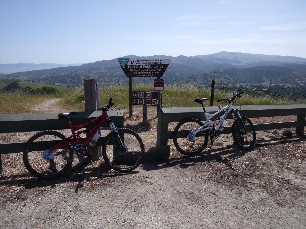 Two mountain bikes, one red and one white, are parked against a green fence at a viewpoint in Fort Ord Public Lands. A sign indicating the park's name and rules is visible in the background, with rolling hills and blue skies framing the scenic view. Fort Ord Public Lands mountain bike trail.