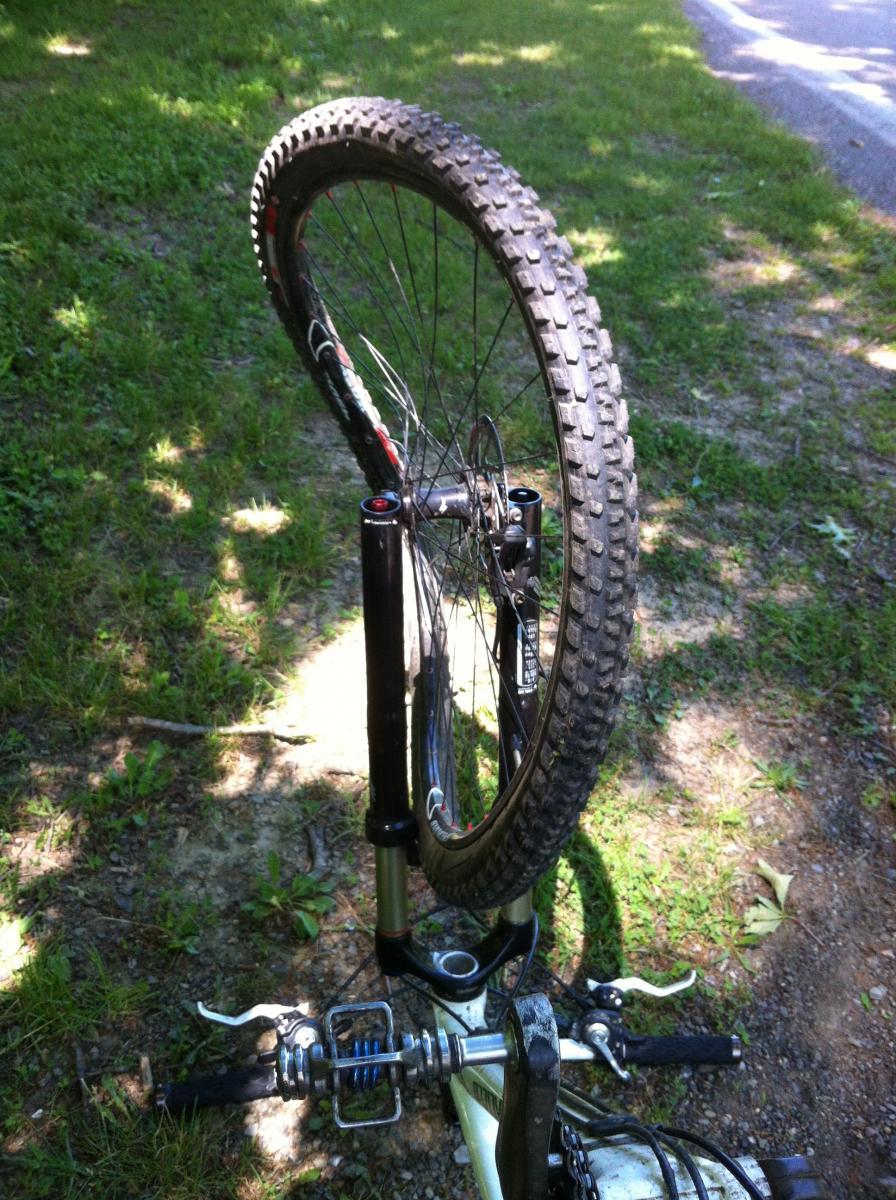 A mountain bike resting on its rear wheel, with the front fork and handlebars visible in the foreground. The setting includes grassy ground and a path in the background, suggesting an outdoor environment. North Park mountain bike trail.