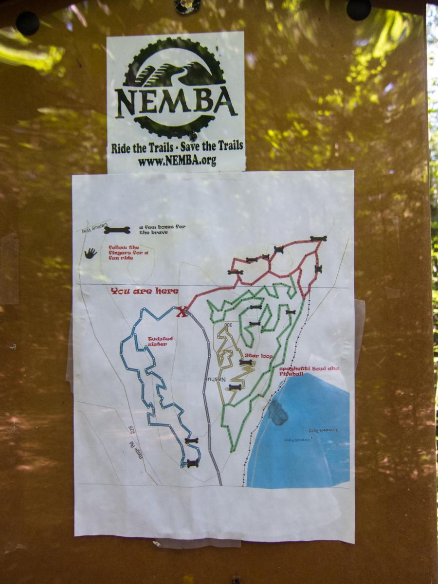 Map showing trails with markings indicating different route options, labeled "You are here" with a location marker. Above the map, there's a sign for NEMBA (New England Mountain Bike Association) promoting trail riding and conservation. The map features various trails in different colors, labeled with names such as "Twisted Disaster" and "Sifter Loop." The background includes a wooden bulletin board. Yudicky Farm mountain bike trail.