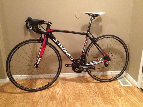Specialized Epic Comp 29er: A black and red S-Works road bike leaning against a wall, featuring thin tires and a lightweight frame, with drop handlebars and a sleek design. The background shows a wooden floor and a neutral-colored wall.