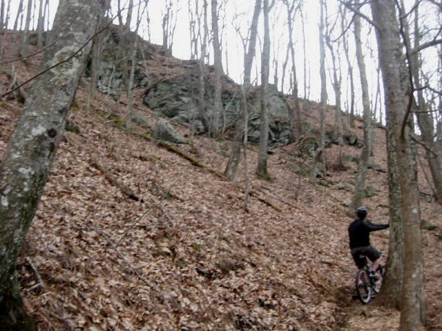 A person riding a mountain bike on a dirt trail in a wooded area, with bare trees and a rocky hillside in the background. The ground is covered with fallen leaves, indicating an autumn or early spring setting. Aska Trail System mountain bike trail.