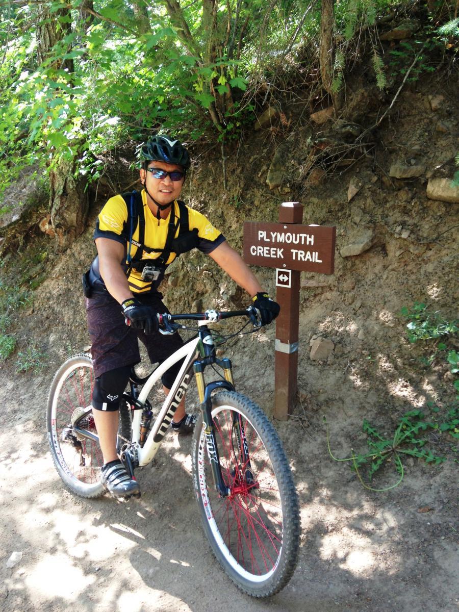 A mountain biker dressed in a yellow and black jersey poses beside a wooden trail sign that reads "Plymouth Creek Trail." The cyclist is wearing a helmet and sunglasses, and their bike is parked on a dirt path surrounded by green foliage and trees. Deer Creek Canyon mountain bike trail.