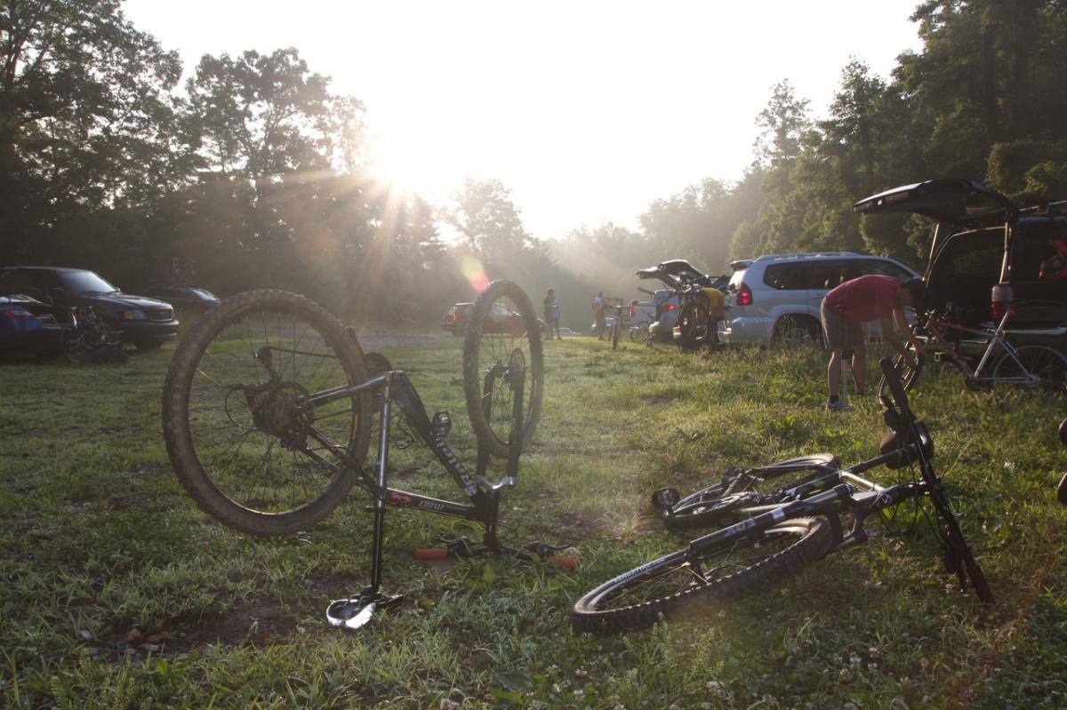A sunrise scene in a grassy parking area with mountain bikes lying on the ground. One bike is propped on its handlebars while another is on its side. In the background, several vehicles are parked, and people are seen preparing for a biking excursion. Sunlight filters through the trees, creating a warm, inviting atmosphere. Bull / Jake Mountain mountain bike trail.