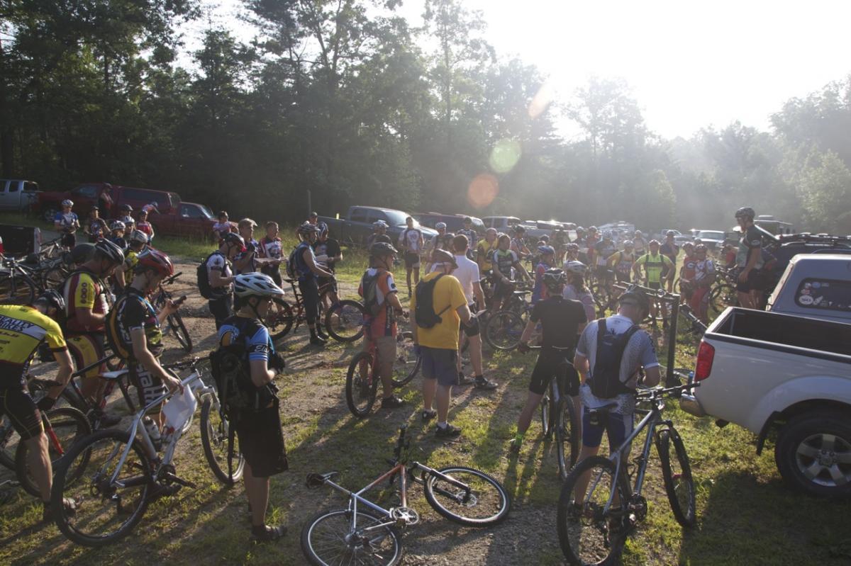 A group of mountain bikers gathered in a wooded area for a pre-ride meeting. Many participants are seen wearing cycling jerseys and helmets, while some are holding their bikes. The setting is early morning with soft sunlight filtering through the trees, and vehicles can be seen parked in the background. Bull / Jake Mountain mountain bike trail.