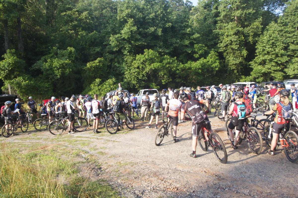 A large group of cyclists gathered at a parking area surrounded by lush greenery. Many participants are wearing cycling gear and helmets, standing with their mountain bikes as they prepare for a ride or event. Several vehicles are parked in the background. The scene captures a vibrant outdoor atmosphere with sunlight filtering through the trees. Bull / Jake Mountain mountain bike trail.