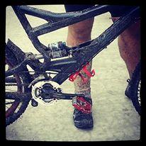 Specialized Enduro Elite: Close-up of a muddy mountain bike with visible drivetrain components, including the pedals and rear derailleur, alongside the muddy lower legs of a person wearing cycling shoes.