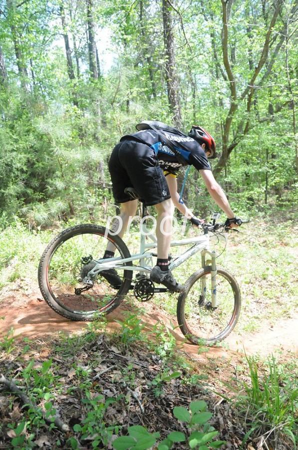 A mountain biker is navigating a dirt path in a lush, green forest. The rider is leaning forward on the bike, wearing a helmet and athletic gear, with one hand on the handlebars. The bike is positioned on a slight incline, surrounded by foliage and trees. Lakeside Trail mountain bike trail.