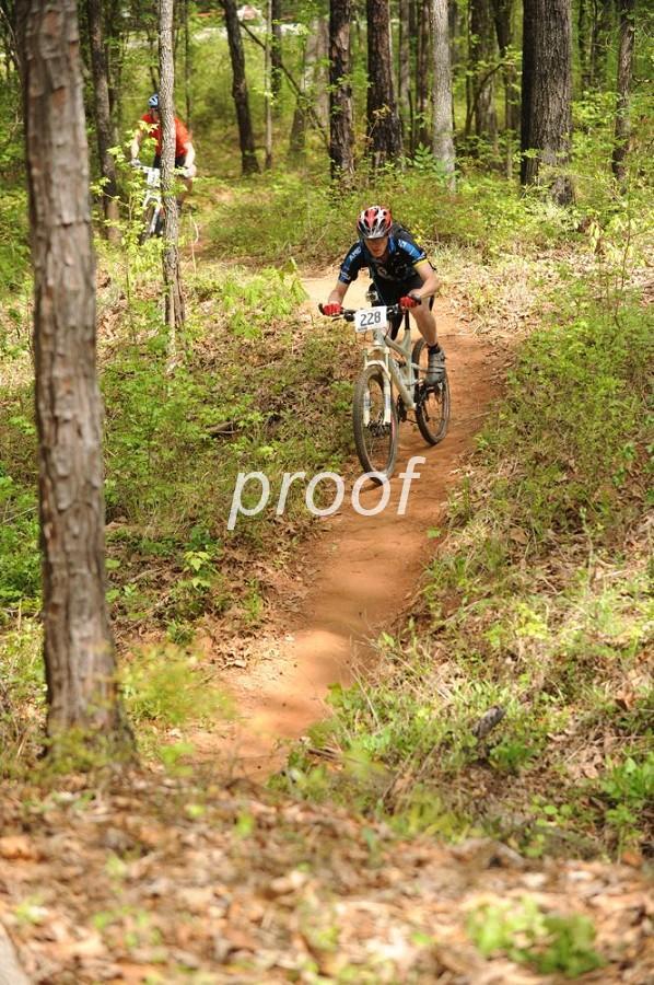 A mountain biker navigating a dirt trail surrounded by trees in a forested area. The biker is wearing a helmet and cycling gear, with a race number visible on their shirt. Another cyclist can be seen in the background, also on the trail. The scene captures the excitement of outdoor sports in nature. Lakeside Trail mountain bike trail.