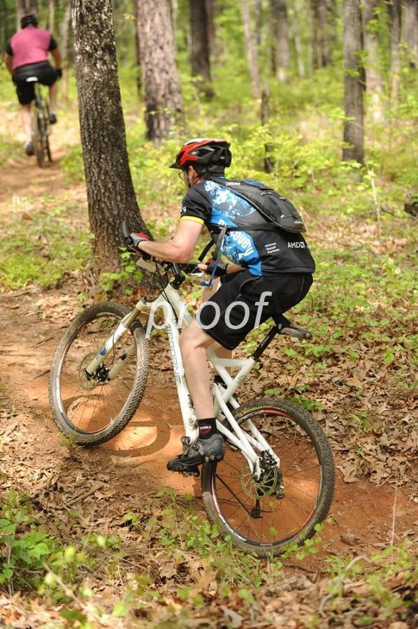 A mountain biker navigating a dirt trail through a wooded area, focusing on maintaining balance while riding. The biker is wearing a helmet and cycling kit, with another cyclist visible in the background. The terrain features trees and green foliage, showcasing a natural outdoor setting. Lakeside Trail mountain bike trail.