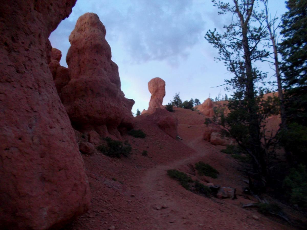 A winding trail leads through a rocky landscape featuring tall, reddish rock formations under a twilight sky. Sparse vegetation can be seen along the path, which is bordered by trees in the background. Thunder Mountain mountain bike trail.