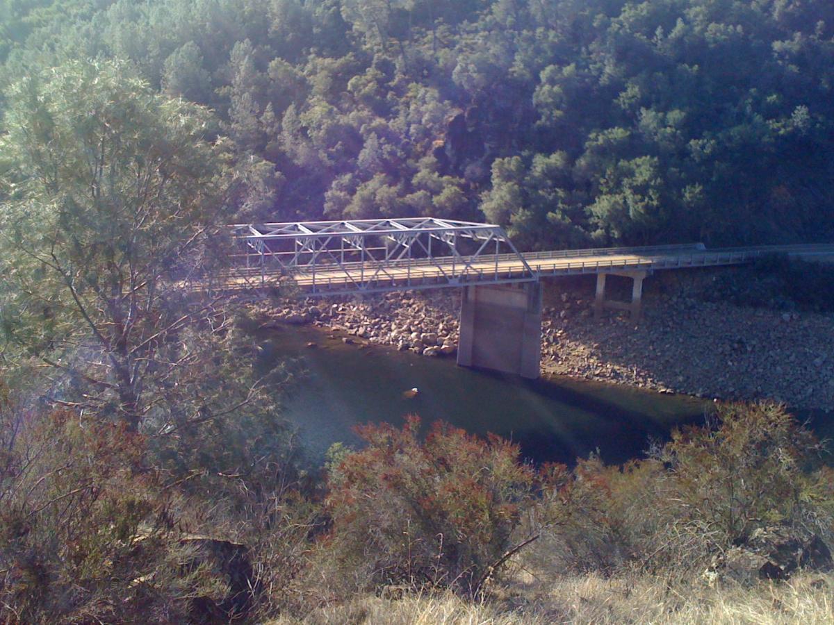 A metal arch bridge spans a river surrounded by greenery. The bridge features a concrete support on one side and is set against a backdrop of hills. Sunlight filters through the trees, creating a natural and serene atmosphere. Sweetwater Trail mountain bike trail.