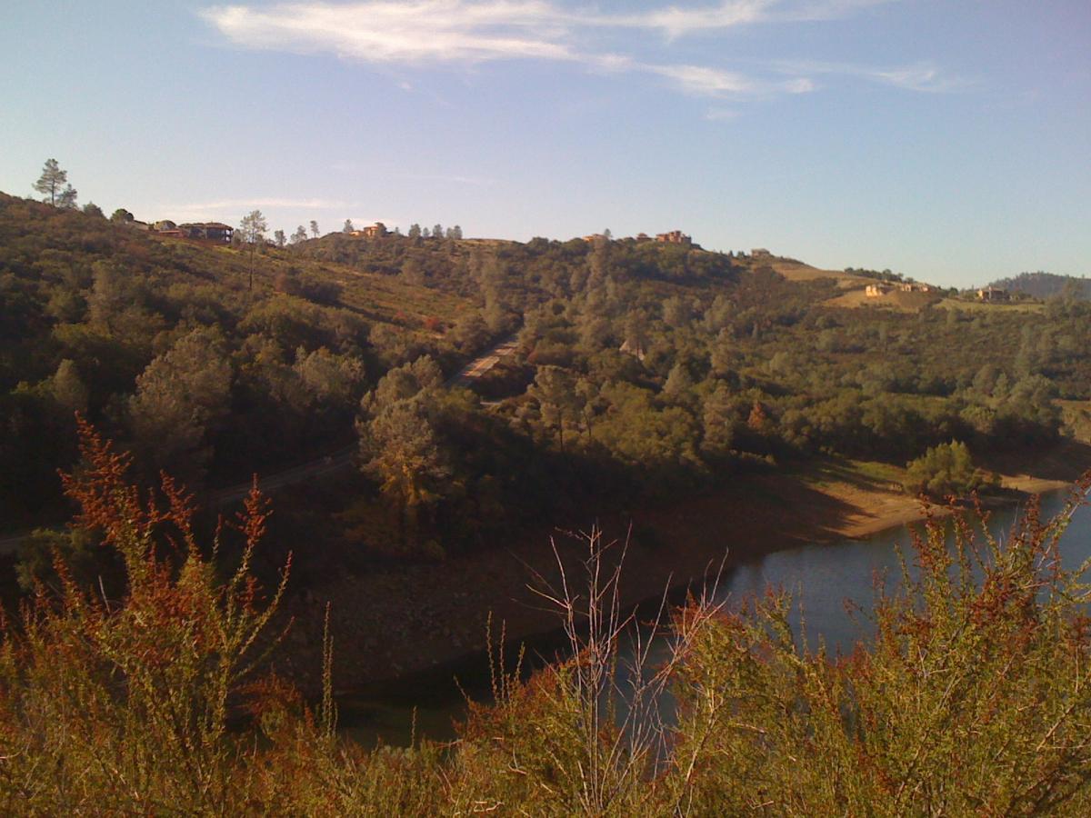 Scenic view of rolling hills covered with greenery, a winding road visible, and a calm body of water in the foreground. Blue sky with some wispy clouds can be seen above, while houses are situated on the hills in the distance. Sweetwater Trail mountain bike trail.