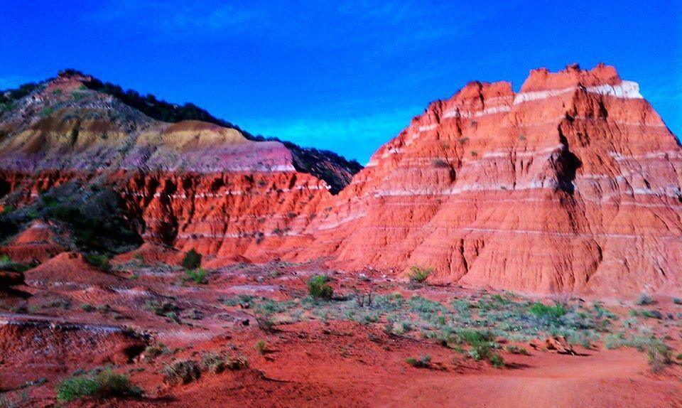 Scenic view of red rock formations with layered textures under a clear blue sky, surrounded by sparse vegetation. The landscape features steep cliffs and vibrant earthy colors, typical of a natural canyon or geological formation. Palo Duro Canyon mountain bike trail.