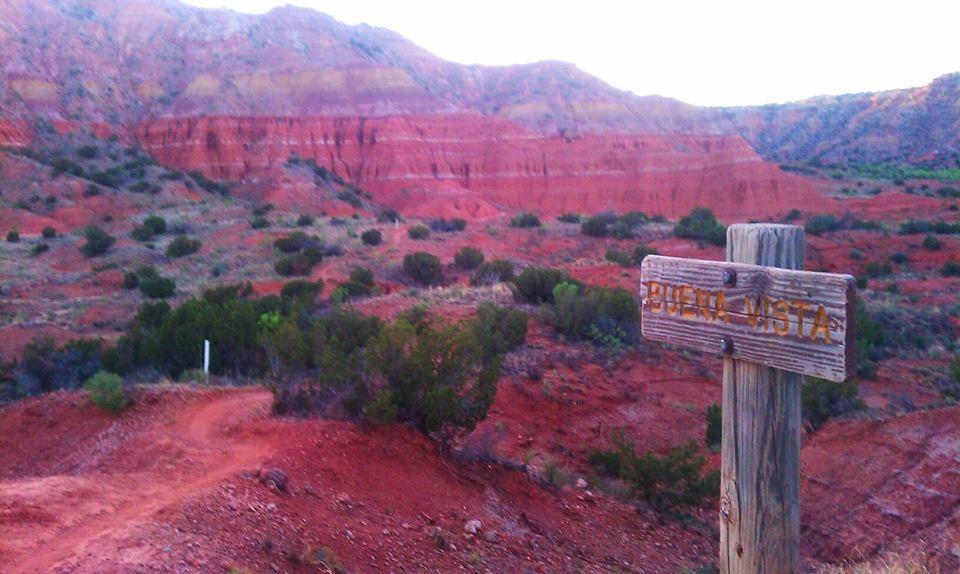 A scenic view of colorful red rock formations and hills, with a wooden sign labeled "Puenta Vista" in the foreground. A dirt path meanders through the landscape, surrounded by sparse vegetation and shrubs under a clear sky. Palo Duro Canyon mountain bike trail.