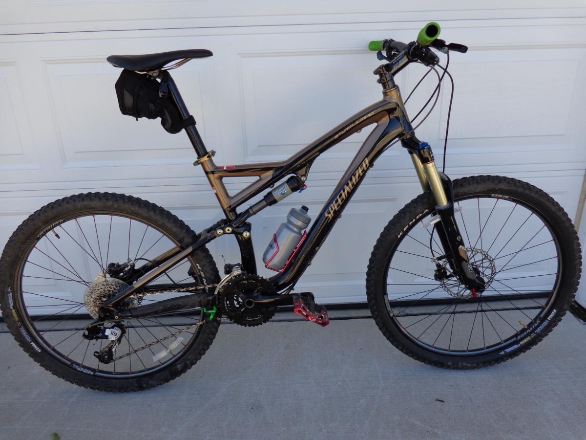 Specialized Stumpjumper FSR: A side view of a mountain bike with a dark frame, featuring a suspension system and thick tires. The bike is equipped with a water bottle and a small black bag attached to the seat post. The background shows a white garage door.