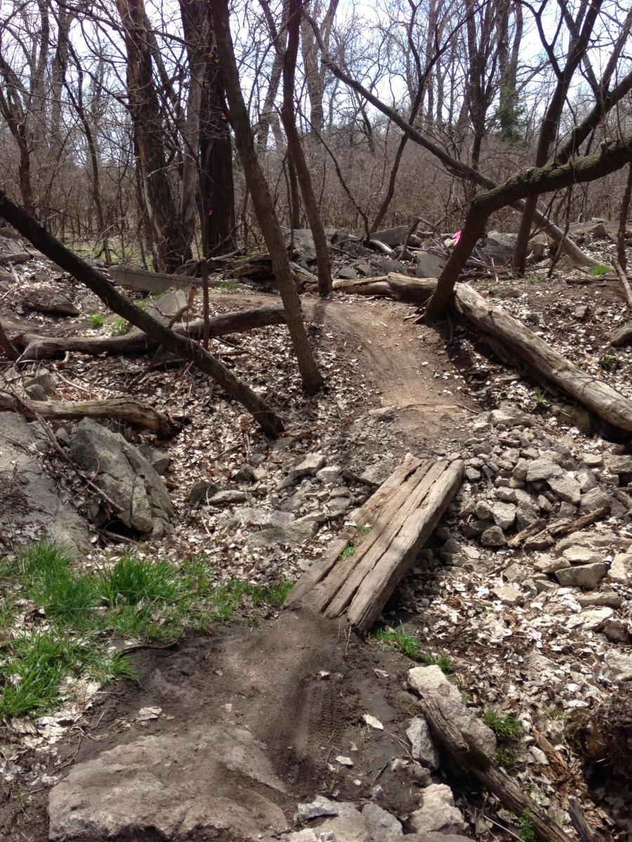A narrow dirt path winding through a wooded area, surrounded by trees and scattered rocks. The ground is covered with fallen leaves, and wooden planks are placed across parts of the path, likely serving as a makeshift bridge. The scene appears to be part of an outdoor trail or biking path in a natural setting. Air Capital Memorial Park mountain bike trail.